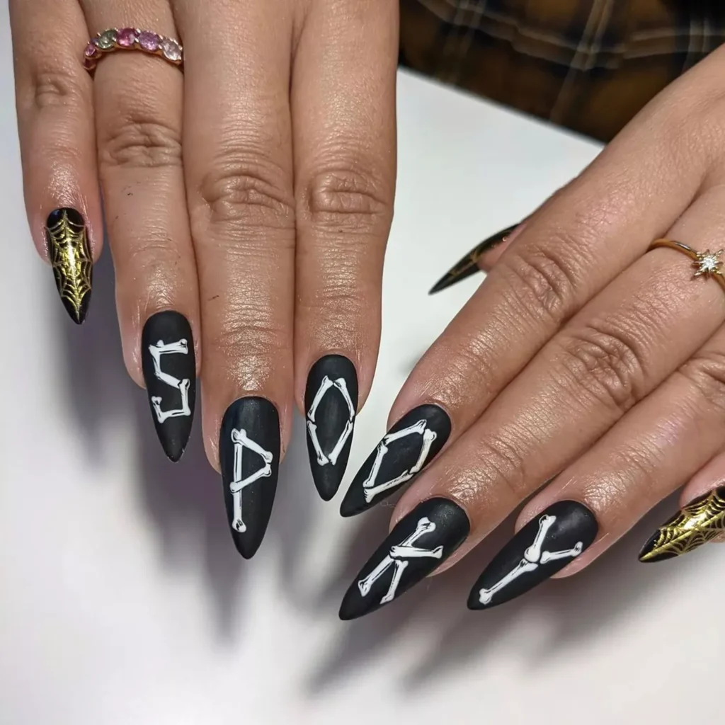 Matte black halloween nails with gold spiderwebs and white bone-letter art on stiletto nails.